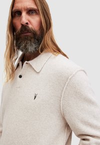 Pullover in maglia beige chiaro con colletto, dotato di chiusura a tre bottoni e piccolo design ricamato sul petto. Tessuto morbido e strutturato.