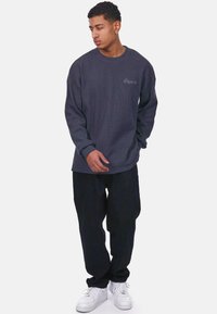 Camisa de manga larga de color gris oscuro con textura acanalada, que presenta un pequeño logo bordado, combinada con pantalones negros holgados y zapatillas blancas.