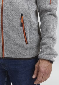 Grå fleecejakke med strukturert stoff, med oransje glidelåsdetaljer, to glidelåslommer og svart kant nederst og ved mansjettene.