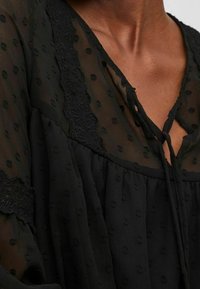 Černá průsvitná halenka s texturovanými puntíky, krajkovými akcenty podél výstřihu a zavazováním na mašli. Měkká, splývavá látka.