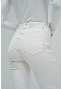Jeans de mezclilla blancos con un diseño ajustado, que cuentan con bolsillos traseros y una etiqueta texturizada en la cinturilla. Tela suave visible.