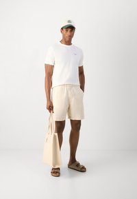 White short-sleeve t-shirt, beige linen shorts, brown slide sandals. Carrying a light beige tote bag. Baseball cap with green brim.