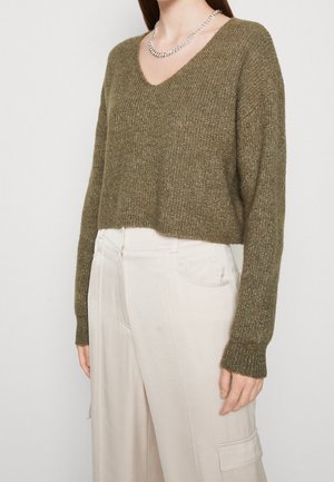 Femme portant un pull court à col en V en maille vert olive et un pantalon blanc ample taille haute avec poches latérales.