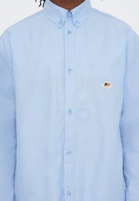 Light blue button-up shirt with a small embroidered logo featuring two cherubs on the left chest, worn by a person.