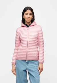 Γυναίκα που φοράει ανοιχτό ροζ μπουφάν με κουκούλα και ανάγλυφο σχέδιο και ανοιχτό μπλε τζιν, όρθια μπροστά σε απλό λευκό φόντο.