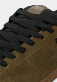 Καφέ σουέτ αθλητικά παπούτσια με μαύρες κορδόνες, με δομημένο πάνελ με διατρήσεις και ελαστική σόλα. Λογότυπο μάρκας στη γλώσσα.