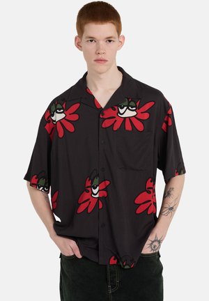 Młody mężczyzna z krótkimi rudymi włosami, ubrany w czarną koszulę na guziki z krótkim rękawem, ozdobioną czerwono-zielonymi abstrakcyjnymi kwiatowymi wzorami, ręce w kieszeniach.