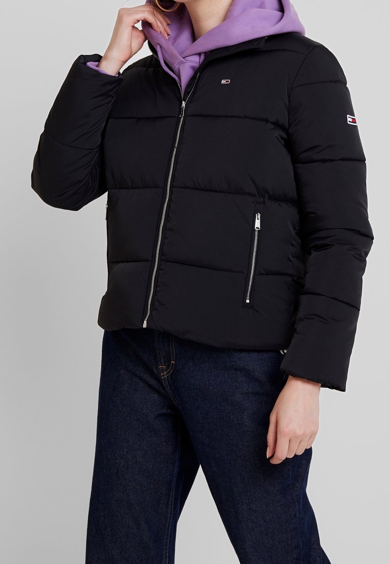 Svart puffjacka med kort design, med dragkedjeförsedda sidofickor, hög krage och vadderad textur, lager ovanför en lila huvtröja.