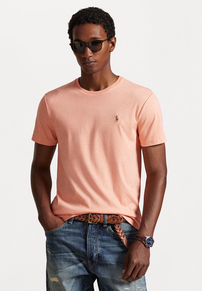 T-shirt à manches courtes en corail clair, fabriqué en tissu doux avec un petit logo brodé sur la poitrine, associé à un jean et des accessoires.