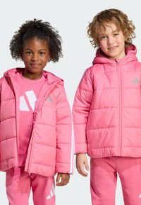 Two children stand side by side, both wearing pink puffer jackets and matching outfits, smiling against a light gray background.