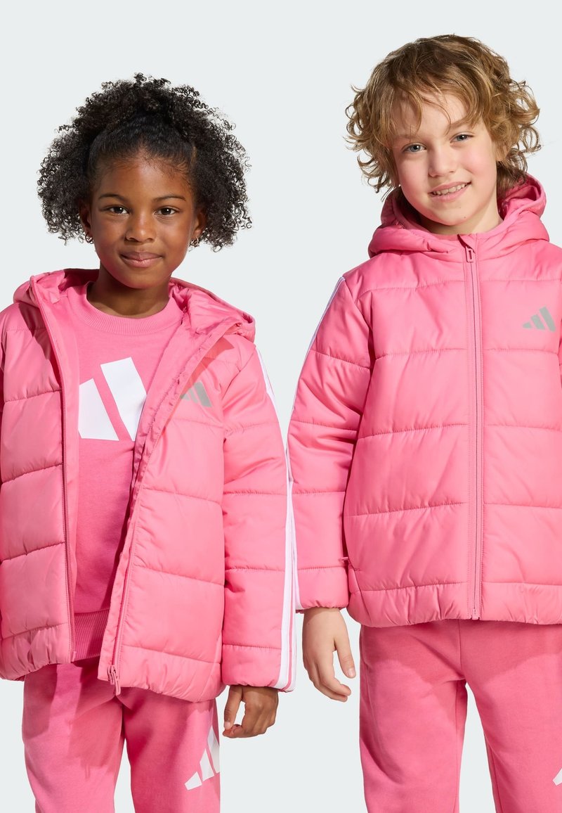 Two children stand side by side, both wearing pink puffer jackets and matching outfits, smiling against a light gray background.