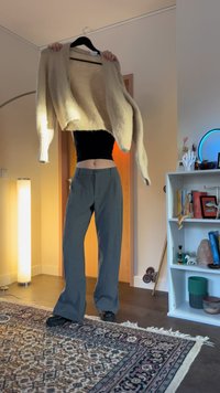 Egy személy bézs kötött pulóvert tart vállfán a feje felett egy mintás szőnyeggel, állólámpával, gördeszkával és fehér könyvespolccal berendezett szobában.