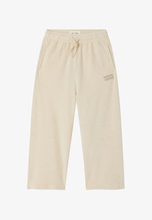 Mladé dieťa so svetlými vlasmi, oblečené v bielom tričku s dlhými rukávmi, béžových širokých nohaviciach a bielych tenisových topánkach, stojace pred bielym pozadím.