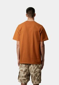 T-shirt di cotone arancione con maniche corte, vestibilità classica e retro semplice, abbinato a pantaloni corti beige a motivo mimetico con tasche laterali.