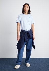Lyseblåt bomulds t-shirt med et blåt logo, mørkeblåt jakke knyttet i livet, løstsiddende blå jeans og cremefarvede sneakers.