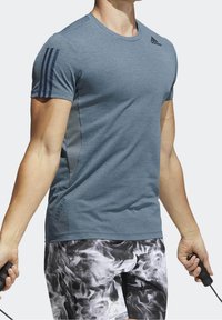 Atleta con camiseta texturizada azul de Adidas y pantalones cortos con patrón blanco y negro sosteniendo las asas de una cuerda para saltar, en una pose intermedia de ejercicio.