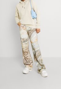 Beige huvtröja, mönstrade vida byxor med virvlar av brunt, kräm och orange, ihop med vita sneakers och en blå handväska.