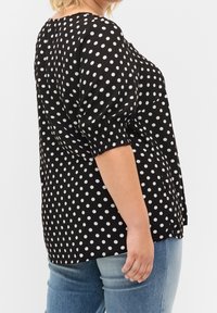 Svart blus med prickar, puffärmar, samlade manschetter och avslappnad passform, i kombination med ljusblå jeans.