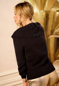 Pulover negru cu glugă ondulată, mâneci texturate și manșete cu nervuri, asortat cu pantaloni aurii lucioși, într-un decor elegant, iluminat suav.