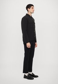 Jachetă neagră din material cu două buzunare frontale, închidere cu nasturi și un croi ajustat. Asortată cu pantaloni negri și pantofi loafers strălucitori negri.