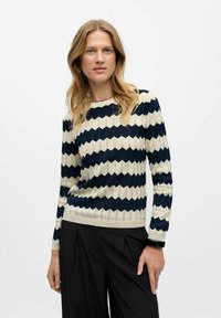 Navy und creme gestreifter Pullover mit geripptem Textur und Wellenmuster. Runder Ausschnitt und lange Ärmel. Kombiniert mit schwarzen Hosen.