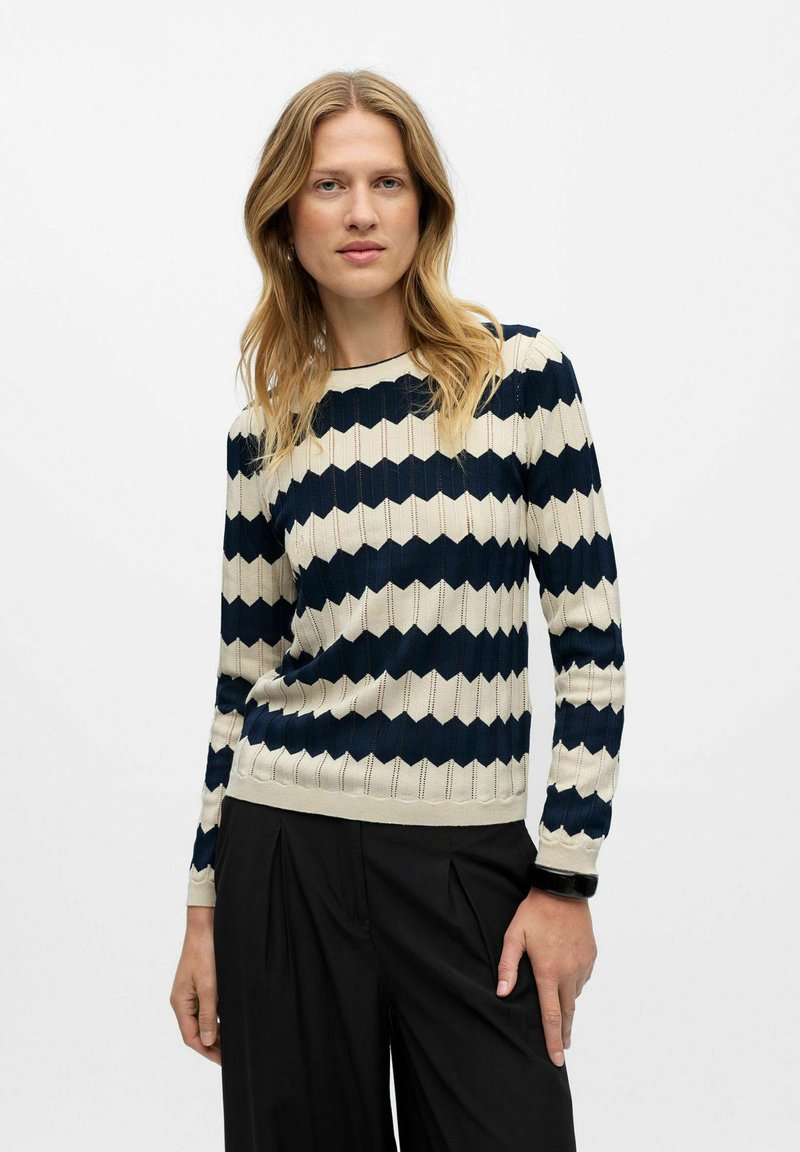 Navy und creme gestreifter Pullover mit geripptem Textur und Wellenmuster. Runder Ausschnitt und lange Ärmel. Kombiniert mit schwarzen Hosen.