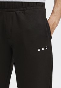 Sorte sweatpants lavet af blødt stof, med elastisk talje og sidelommer. Det højre ben har "A.R.C." trykt i hvidt.