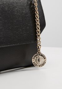 Borsa a mano in ecopelle nera texturizzata con tracolla a catena dorata e targhetta rotonda in oro con incisione "Donna Karan 1988".