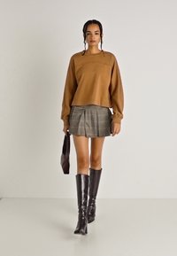 Top lung cu maneci lungi de culoare maronie si fusta mini plisata gri cu carouri, asortate cu cizme lucioase maro pana la genunchi si o geanta asortata.