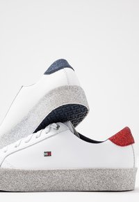 Baskets basses blanches avec semelles à plateforme argentée scintillante, talon bleu marine pailleté sur une chaussure, talon rouge pailleté sur l’autre, et petit logo sur les côtés.