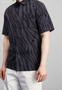 Košile s krátkým rukávem v tmavě modré barvě s černým vzorem zebřích pruhů. Má zapínání na knoflíky vpředu a náprsní kapsu. Vyrobena z lehké látky.