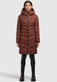 Braune Pufferjacke mit Chevron-Design, Reißverschluss vorne, verstellbarer Kapuze und Seitentaschen, kombiniert mit schwarzen, figurbetonten Hosen und Stiefeln.