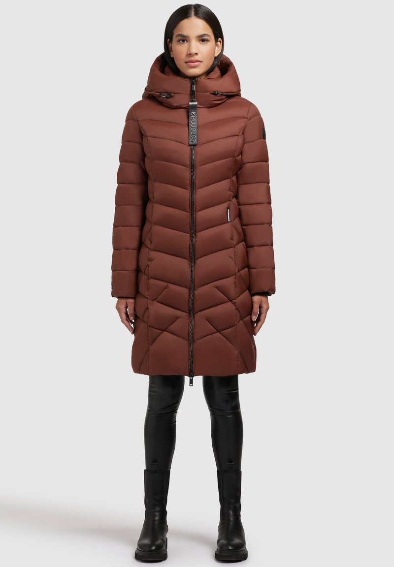 Braune Pufferjacke mit Chevron-Design, Reißverschluss vorne, verstellbarer Kapuze und Seitentaschen, kombiniert mit schwarzen, figurbetonten Hosen und Stiefeln.