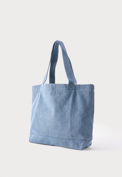 Sac fourre-tout en denim bleu avec deux bretelles, tenu debout contre un fond clair.