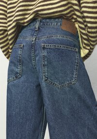 Detailný záber na osobu v modrých rifliach a prúžkovanom hnedom a béžovom svetri, so zameraním na oblasť zadných vreciek.