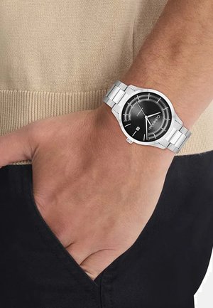 Montre en acier inoxydable argenté avec un cadran noir, des index argentés et un affichage de la date. Portée au poignet, avec un bracelet à maillons.