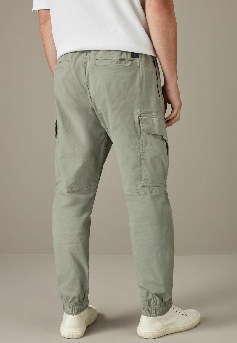 Homme portant un pantalon cargo vert clair avec des bords élastiques et des baskets blanches, debout devant un fond beige uni.