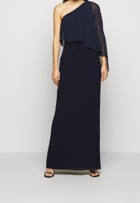 Navyblå klänning med en axel och ett lager av chiffong på ena sidan, golvlång design och åtsittande liv. Enkla svarta sandaler med remmar.