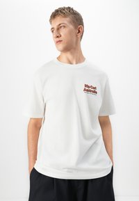 Ung mann med blondt hår iført hvit Rip Curl Australia t-skjorte og svarte shorts, ser mot venstre med hendene i lommene.
