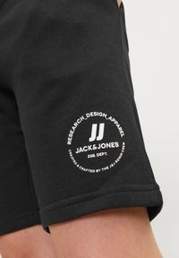 Zwarte katoenen shorts met een rond wit logo met tekst op de linkerdij. Het ontwerp heeft een zichtbare zoom en een zijzak.