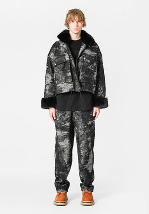 Joven con chaqueta texturizada negra y gris con cuello y puños de piel, pantalones a juego, camiseta negra y zapatos marrones, de pie en posición recta.