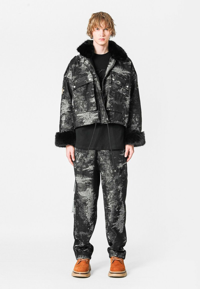 Joven con chaqueta texturizada negra y gris con cuello y puños de piel, pantalones a juego, camiseta negra y zapatos marrones, de pie en posición recta.