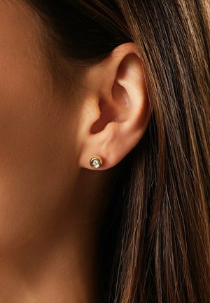 Goldene Steckerohrringe mit einem klaren, runden Edelstein in der Mitte, positioniert im Ohrläppchen, vor langen braunen Haaren.