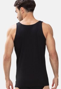 Mann mit kurzem Haar, der ein ärmelloses schwarzes Tank-Top trägt, mit entspannten Armen an den Seiten von einem einfarbigen Hintergrund abgewandt.