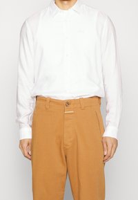 Vit långärmad skjorta med krage, mjukt tyg och subtil brodering, i kombination med bruna byxor med knapp och sömdekor.