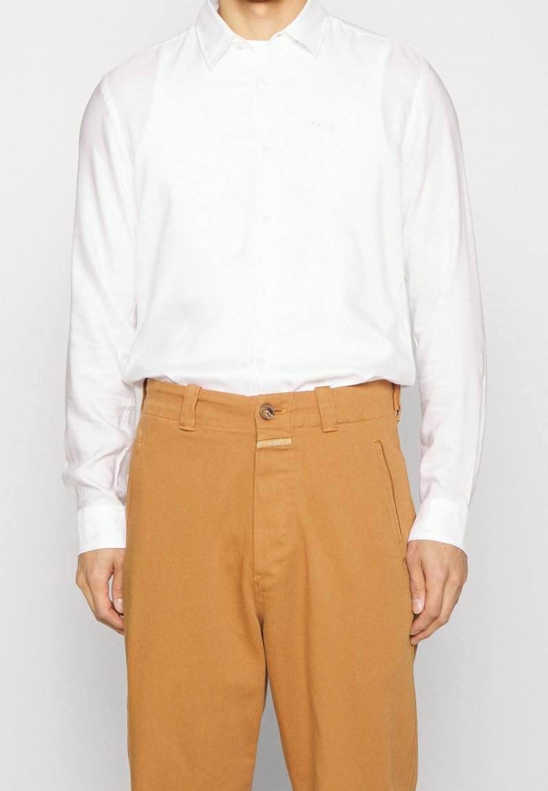 Vit långärmad skjorta med krage, mjukt tyg och subtil brodering, i kombination med bruna byxor med knapp och sömdekor.