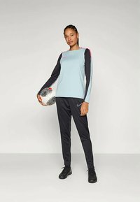 Gaiši zils garām piedurknēm sporta tops ar tumšām piedurknēm un rozā akcentiem, apvienots ar melnām biksēm. Melnais futbola bumba turēta vienā rokā.