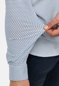 Weißes Hemd mit einem blauen dreieckigen Muster, ausgestattet mit einem knöpfbaren Manschette und glatter Textur. Der Stoff ist leicht und am Handgelenk tailliert.