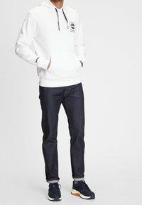 Man i vit Timberland-hoodie, mörkt uppvikt jeanstyg, grå strumpor och marinblå sneakers som står med händerna i hoodie-fickorna.