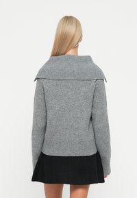 Pull en laine gris avec un large col, des manches longues et des bordures côtelées. Le dos présente un design texturé, superposé à une jupe noire.